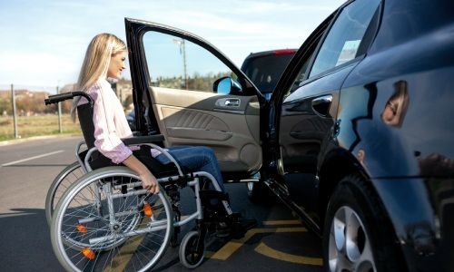 Person, die einen Rollstuhl benutzt wartet vor der offenen Tür eines PKW